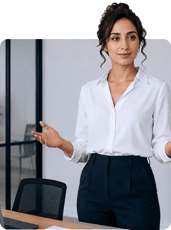 A woman with dark hair in an updo, wearing a white blouse and dark trousers, stands next to a desk with her hands slightly raised, appearing confident in a modern office setting.