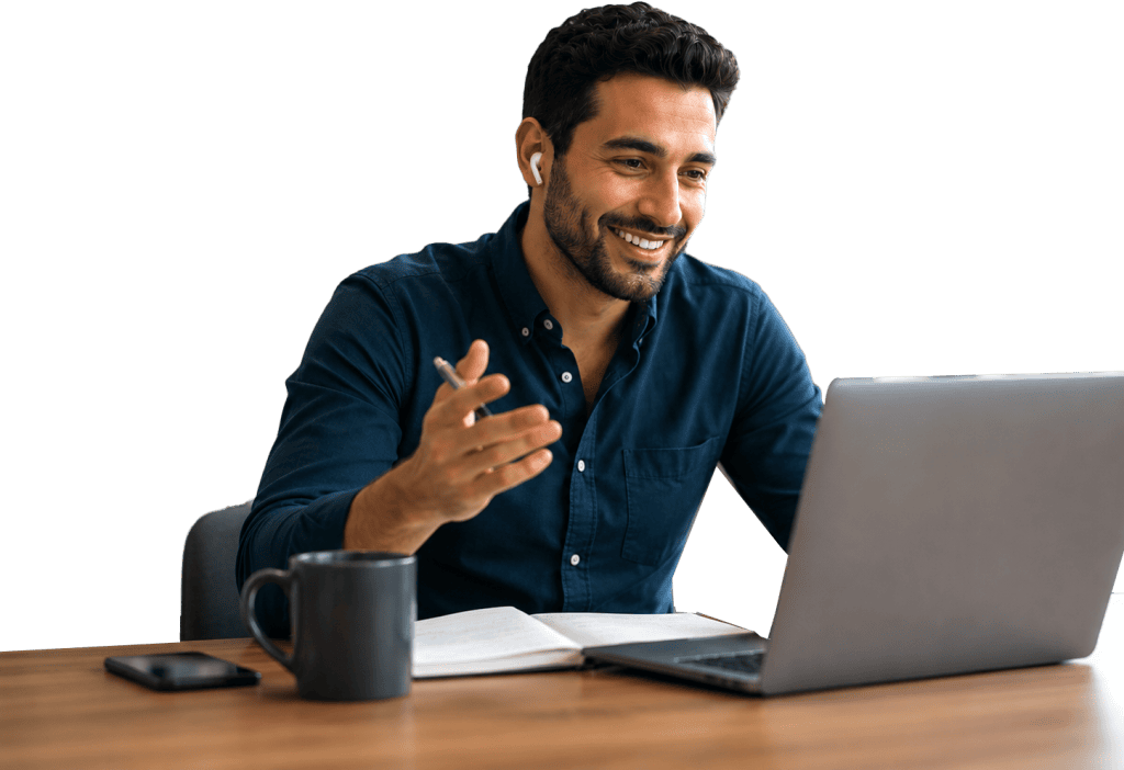 Man wearing earbuds smiles and gestures while video chatting on a laptop at a desk, with a notebook, mug, and smartphone beside him.