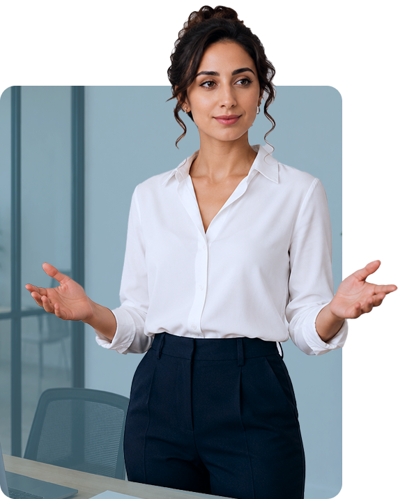 A woman with dark curly hair, wearing a white blouse and navy pants, stands in an office setting, gesturing with both hands as if speaking or presenting.