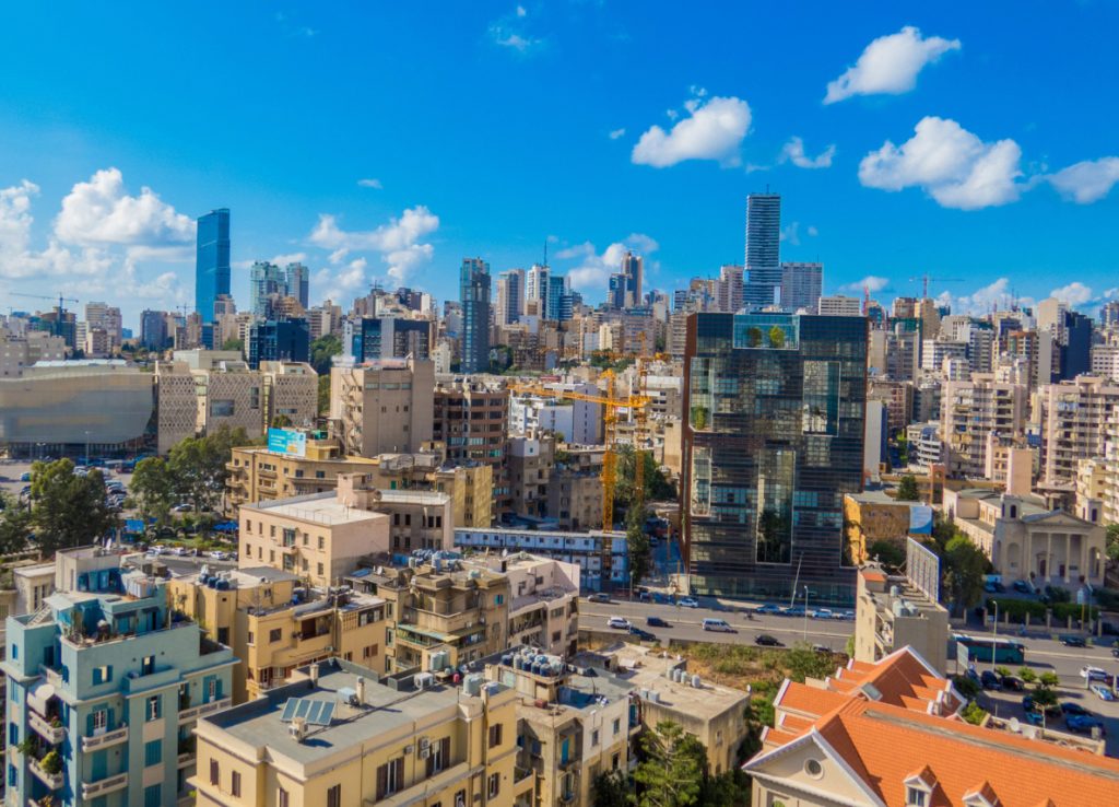 A cityscape of Beirut, Lebanon, featuring a mix of modern skyscrapers and older buildings under a bright blue sky with scattered white clouds.
