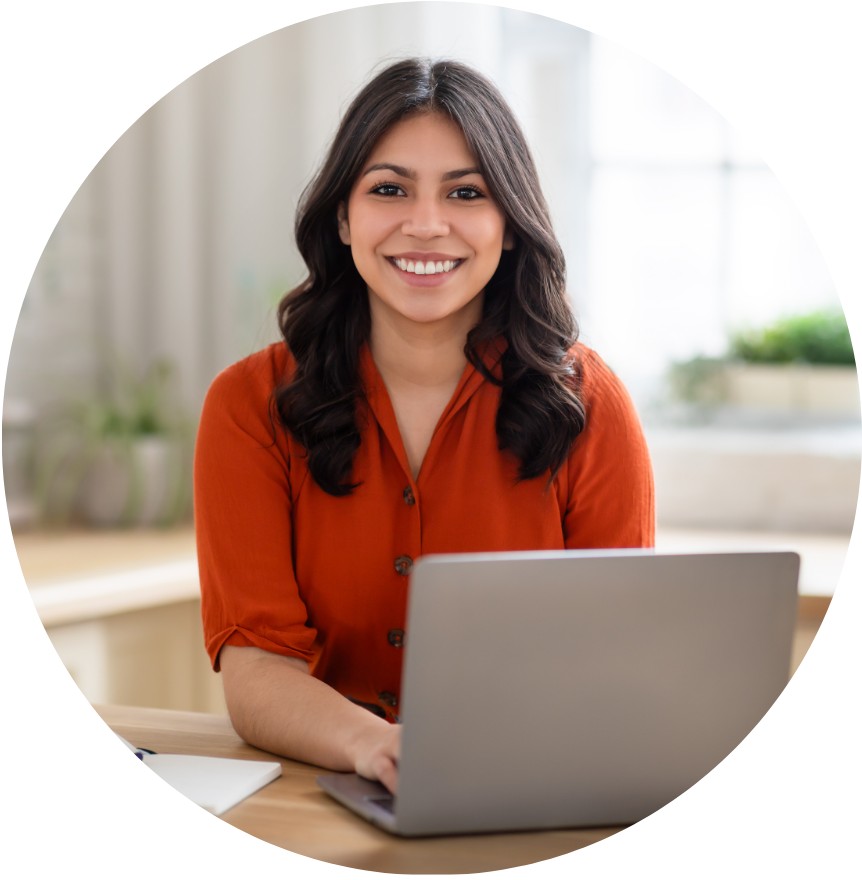 Smiling woman with long dark hair, wearing an orange blouse, sits at a desk using a laptop in a brightly lit, modern home office space with plants in the background.